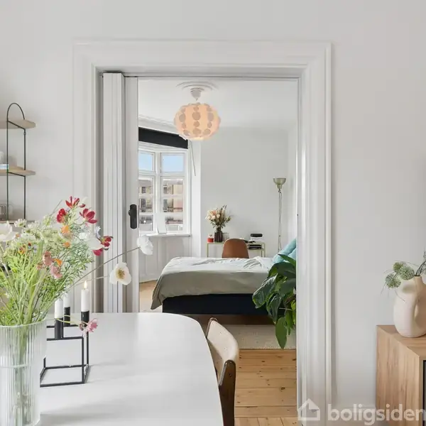 Et moderne interiør med blomster i en vase på et spisebord i forgrunden. Bagved er der adgang til et soveværelse gennem åbne hvide døre, med naturligt lysindfald.