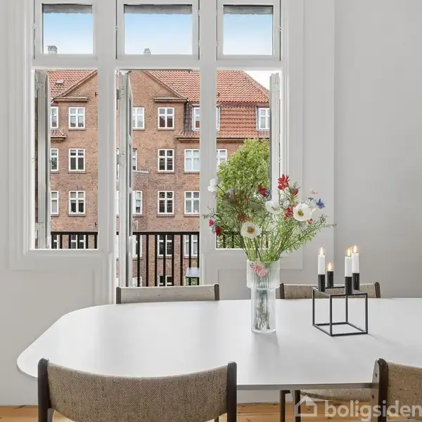 Spisebord med blomster i vase og lysestage med tændte stearinlys står foran et vindue med udsigt til røde murstensbygninger. Et maleri hænger til venstre, hylder til højre.