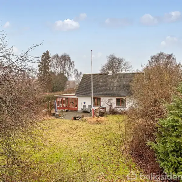 Et hvidt hus med gråt tag står stilrent i en tætmossig have med græsplæne og buske. En flagstang står foran huset i en landlig omgivelser under en blå himmel.