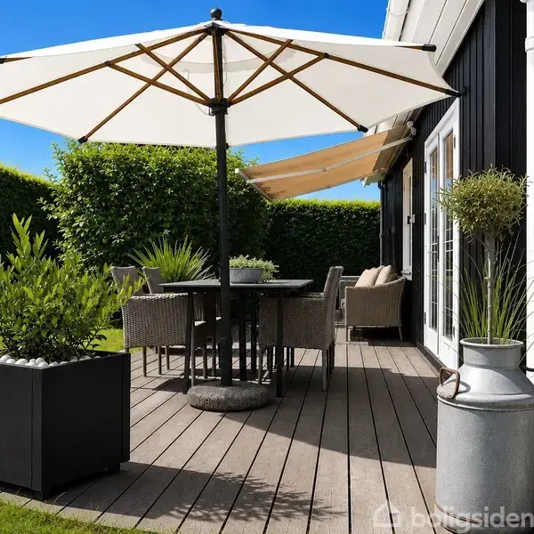En terrasse har et bord og stole under en stor parasol. Omkring er blomsterkasser og en metalbeholder med planter. Græsplænen og hække omkranser terrassen i et solrigt udemiljø.
