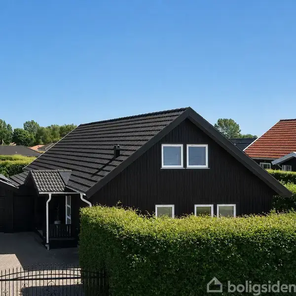 Sortmalet hus med gavl og vinduer, omgivet af grønne hække. Det står i en rolig boligkvarter med blå himmel og nabohuse med røde tage.