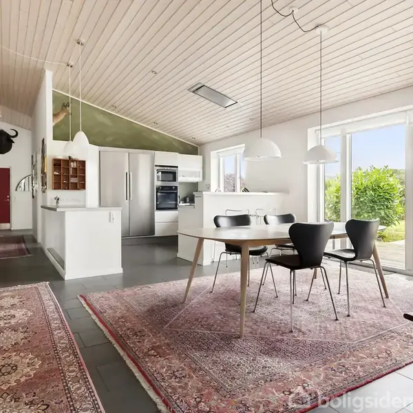 Spisebord med stole står på en rød orientalsk tæppe i et lyst, moderne køkken-alrum. Store vinduer giver udsigt til grøn natur udenfor. Loftslamper hænger over bordet.