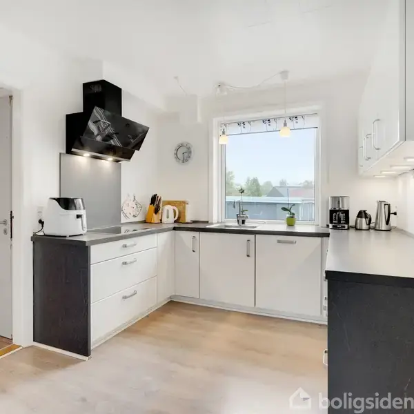 Et moderne køkken med hvide skabe og sort bordplade. En emhætte med skråt design hænger over kogepladen. Et vindue lader naturligt lys ind fra siden.