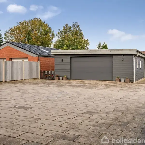 En stor, grå garage med lukket port står på en rummelig flisebelagt gårdsplads, omgivet af et stakit og flere træer i baggrunden.