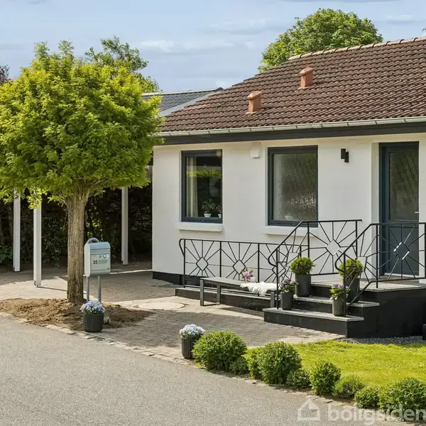 En hvid enfamilieshus med brunt tag står langs en vej. Huset har en terrasse med blomsterkrukker foran, og en carport ligger ved siden af med grønne buske og et lille træ i nærheden.