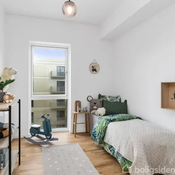 Et børneværelse med en enkeltseng dækket af et grønt tæppe, en træhylde med legetøj, og en legetøjshest på gyngestativ. Et vindue giver udsigt til en bygning udenfor.