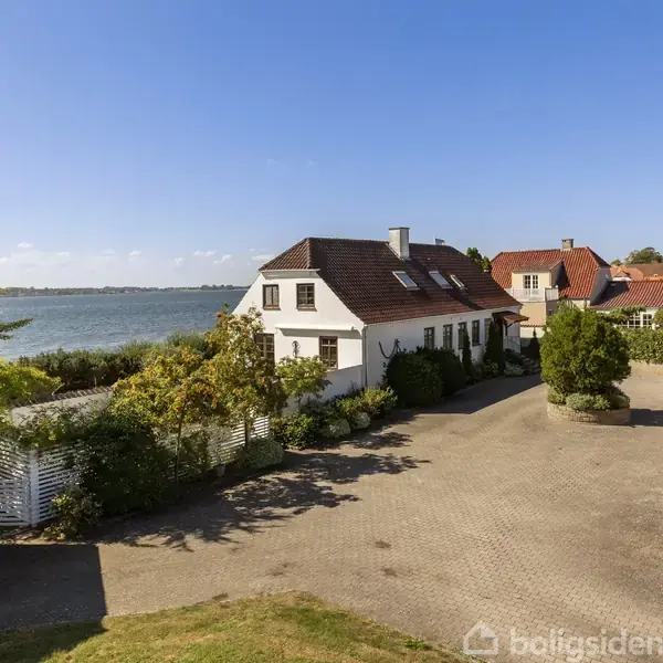 Hus med hvid facade og rødt tag ligger tæt på kysten. Gårdsplads omgivet af grønne planter. I baggrunden ses flere huse og en blå himmel over havet.