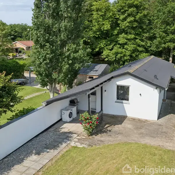 Huset har en hvid facade og sort tag, omgivet af grønne træer og hække. En flisebelagt sti fører til hoveddøren, og der er en luftvarmepumpe på siden.