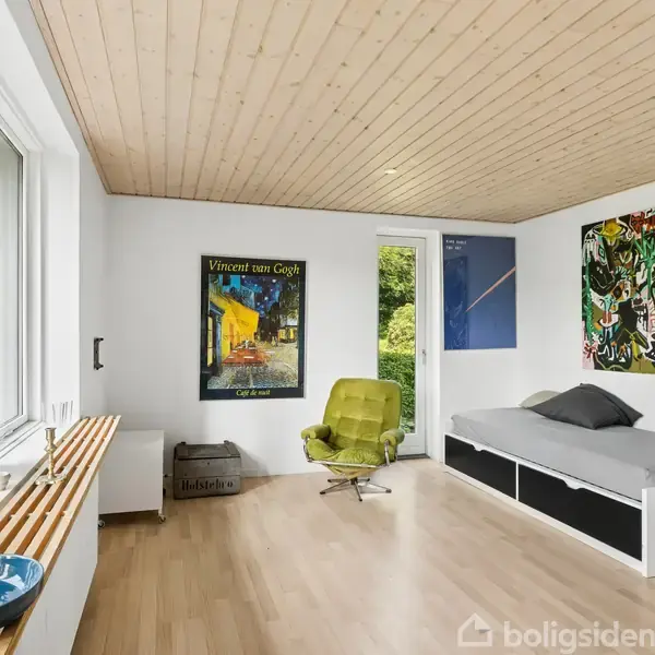 Et værelse med en grøn lænestol står foran en plakat med teksten "Vincent van Gogh". En grå sovesofa er til højre. Store vinduer giver udsigt til grønne planter udenfor.