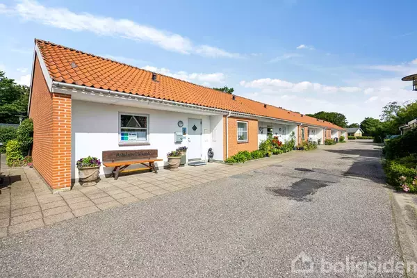 En lang, rød murstensrækkehusbygning med skråt teglstenstag. Foran bygningen står en bænk og to blomsterkrukker. Bygningen er omgivet af en stille villavej og grønne haver.