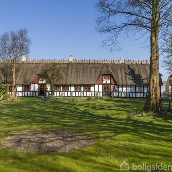 Stråtækt bindingsværkshus står stille i en frodig have med grøn græsplæne, omgivet af træer under en klar blå himmel.