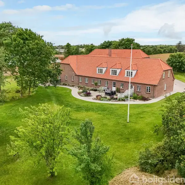Stor, rød teglhúss med rødt tag står stille omgivet af grønne træer og en velplejet græsplæne. En flagstang står i forgrunden under en blå himmel.