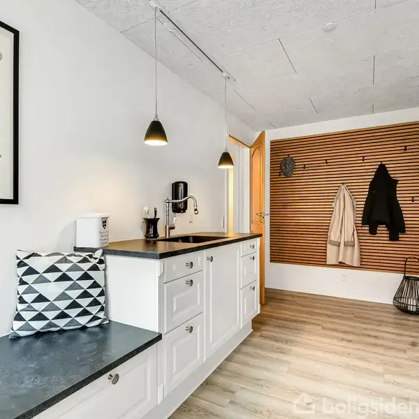 Køkkenet er indrettet med hvide skabe, en mørk bordplade og en håndvask. Væggen har en opslagstavle med hængende jakker og planter. Tekst på en plakat: "Hvis du vil have et smil".