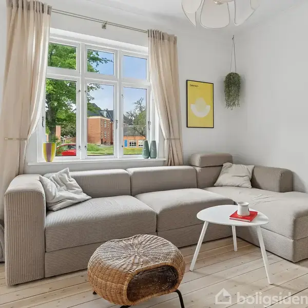 Stor, grå hjørnesofa med puder står ved siden af et lyst vindue med gardiner. Et lille, hvidt bord med kaffekop står foran sofaen. Rummets vægge er hvide.