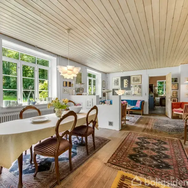 En spisestue med et bord dækket med en lys dug og seks stole. Rummet har trægulv, store vinduer og mange malerier. Et sofaområde ses i baggrunden.