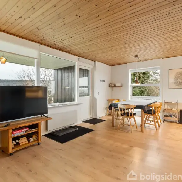 Et rummeligt lyst opholdsområde med trægulv indeholder et TV på et skab og et spisebord med stole. Store vinduer giver udsigt til en have. En lampe hænger fra træloftet.