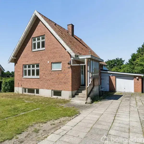 Rødt murstenshus med skråt tag har en lille trappe ved hoveddøren. Omgivet af en græsplæne og flisebelagt indkørsel, står en garage op ad huset. Solrigt, klart vejr.