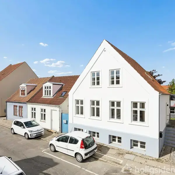Flere hvide huse med røde tage står på en solrig gade. To parkerede biler ses foran dem, med en klar himmel som baggrund.