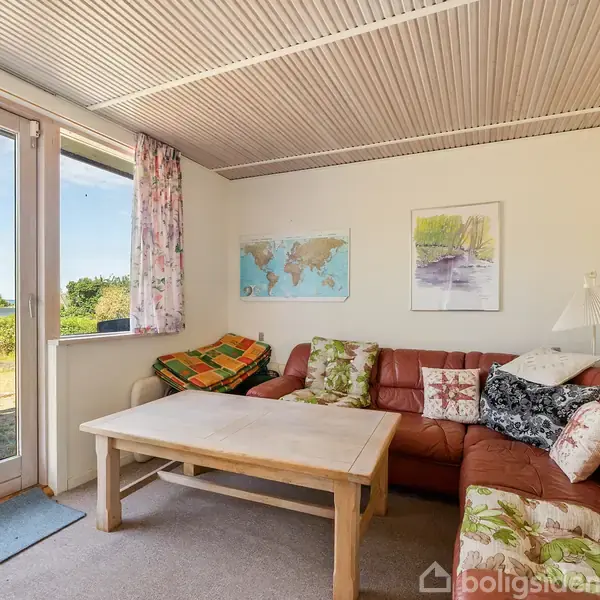 Brun hjørnesofa med forskellige puder står i et rum med træloft. Der er et træbord foran sofaen, vinduer med blomstrede gardiner og natur udenfor. Kort og billede på væggen.