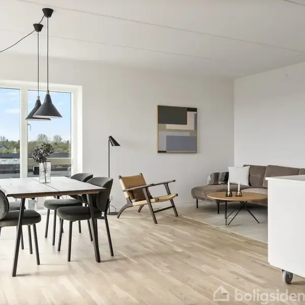 Spisebord med seks stole står i lys stue. En sofa og lænestol supplerer, mens kunst pryder væggen. Store vinduer giver udsigt til grønne omgivelser.