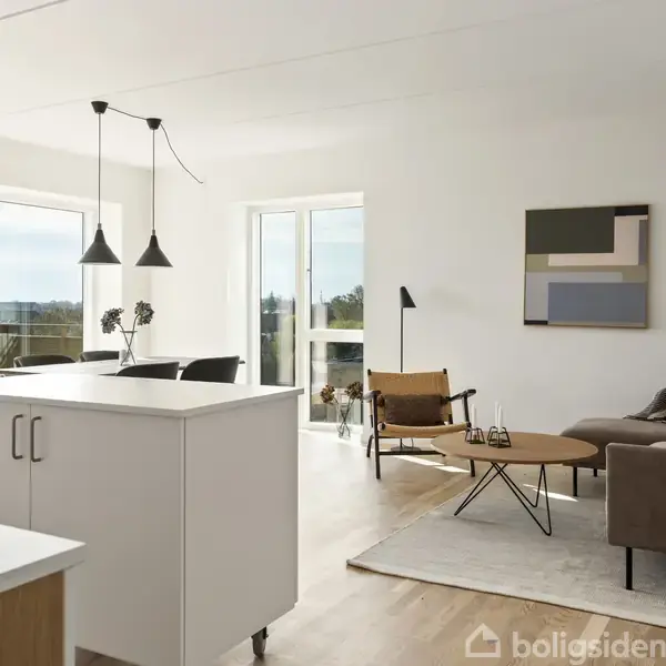 Stue med brun sofa, sofabord på tæppe, moderne loftslamper over en hvid køkkenø. Store vinduer giver udsigt til balkon. Vægkunst hænger over sofaen.