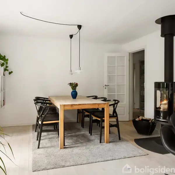 Et spisebord i træ med sorte stole står på et gråt tæppe. En blå vase med blomster pryder bordet. I hjørnet er en moderne brændeovn tændt. En plante står i forgrunden, og der er god belysning fra vinduerne.