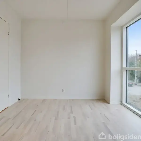 Et tomt rum har lyse trægulve og en stor glasvæg til højre, der giver udsigt til en vej med byggearbejde udenfor. Væggene er hvide og loftet har en enkel loftlampe.