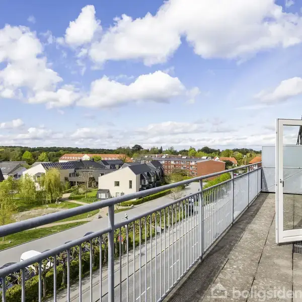 Balkon med gelænder strækker sig langs en bygning, med dør åben til en udsigt over huse og træer i et beboelsesområde under en blå himmel med skyer.