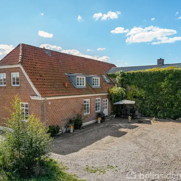 Et murstenshus med rødt tag står stille i en solrig gård omgivet af grønt, buske og et parasolområde ved siden af en vej og andre bygninger.