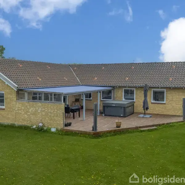 Et gult murstenshus med terrasse og jacuzzi, omgivet af græsplæne og træer under en blå himmel med skyer.