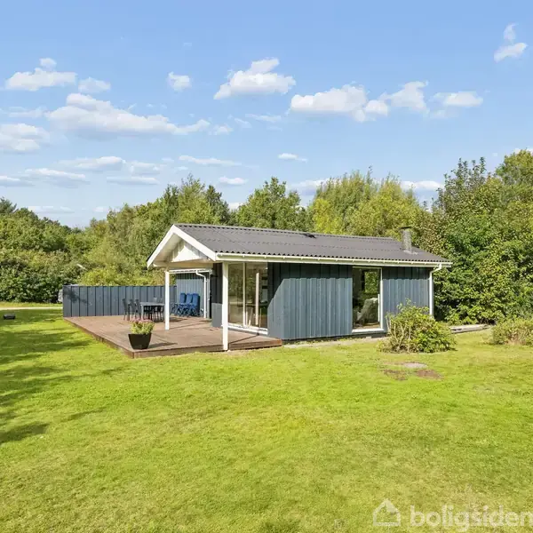 Et hvidt og blåt sommerhus står stille omgivet af en grøn græsplæne og træer, under en blå himmel med hvide skyer.