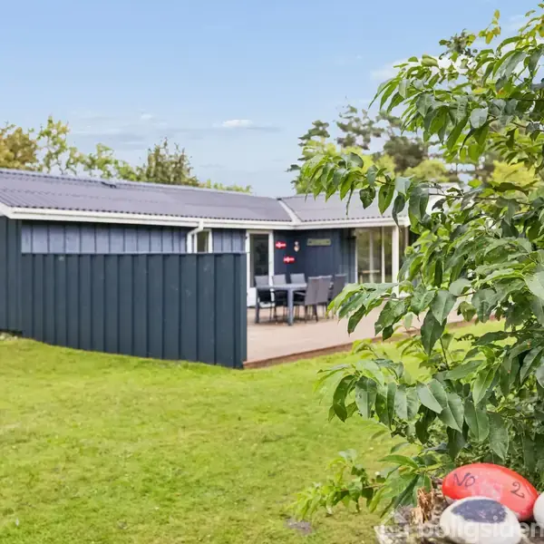 En blå træbygning med terrasse og havemøbler står i en frodig have omkranset af grønne buske og græsplæne. Udsigten viser en blå himmel og grønne træer.