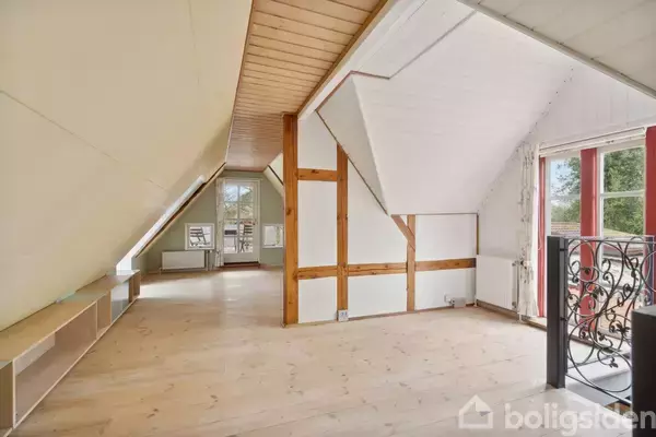 Et lyst, rummeligt loftsrum med træloft og gulv. Der er en indbygget hylde på den ene side og synlige træbjælker. Store vinduer giver udsigt til et grønt område udenfor.