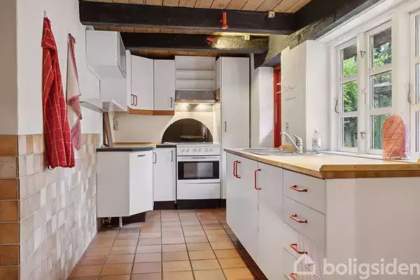 Køkken med hvide skabe og røde greb. Komfuret er placeret centralt. Flisegulv og vægklinker. Et vindue giver udsigt til grønne omgivelser. Røde viskestykker hænger på væggen.