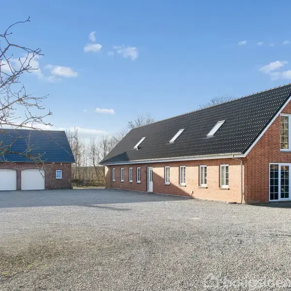 Rødt murstenshus med sort tag, store vinduer, og indgangsdør. Grusbelagt gårdsplads foran, garagebygning til venstre, omgivet af træer under en blå himmel. Ingen tekst fundet.