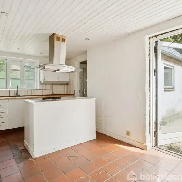 Køkken med hvidt skabsmiljø og central ø, udsmykket med en emhætte. Dør åbner mod udendørs terrasse med fliser, omgivet af grønt. Vandhane og ovn er synlige.