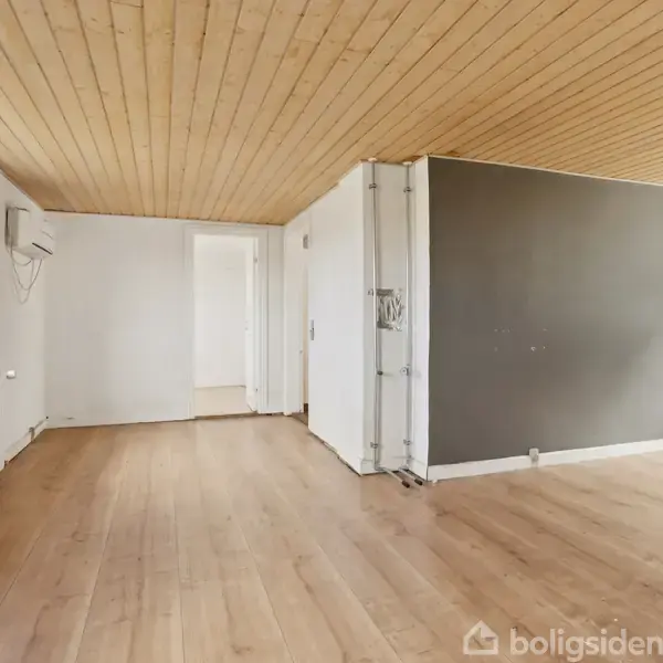 Stue med trægulv og træloft, store vinduer til venstre og en gråmalet væg på bagsiden. Rummet er lyst og åbent, med hvidmalede vægge og et radiator langs vinduet.
