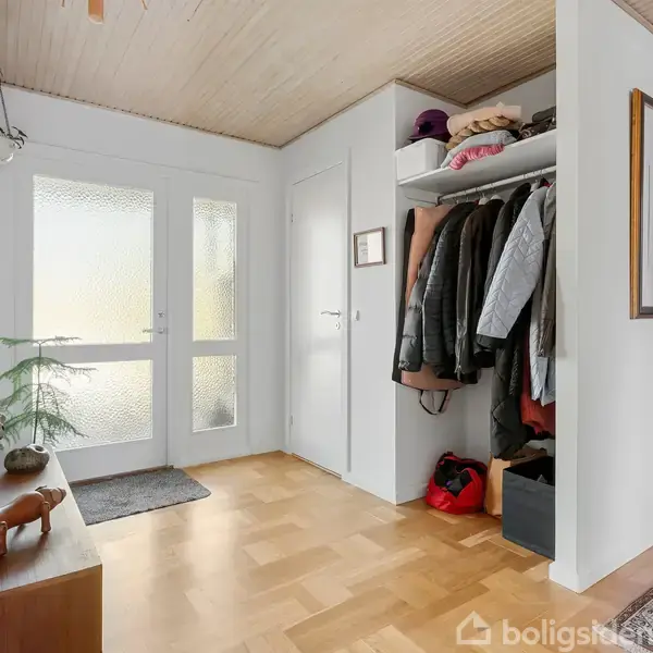Et indgangsparti med en garderobe fyldt med jakker og hatte. En skohylde placeret under tøjet. Daglys trænger ind gennem glasdør, og et lille træ står ved siden af den.