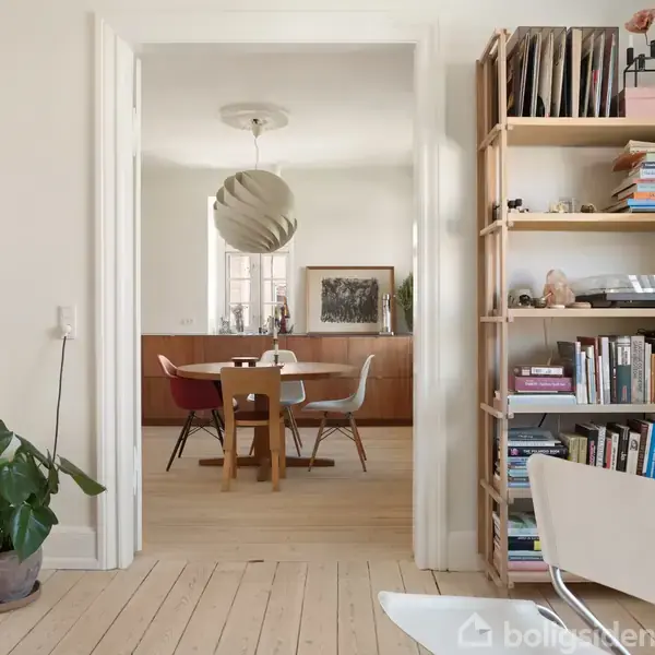 En lys stue med en hylde fyldt med bøger til højre, planter til venstre, og et døråbning fører til spisestue med et bord og stole samt en moderne lampe.