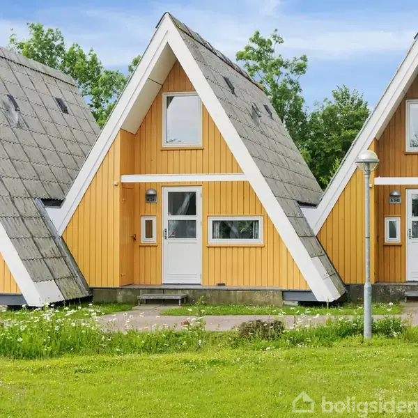 Et gult, A-formet sommerhus står på en grøn græsplæne med andre lignende huse i baggrunden. Huset har hvide vinduer og en dør, omgivet af træer og en blå himmel.