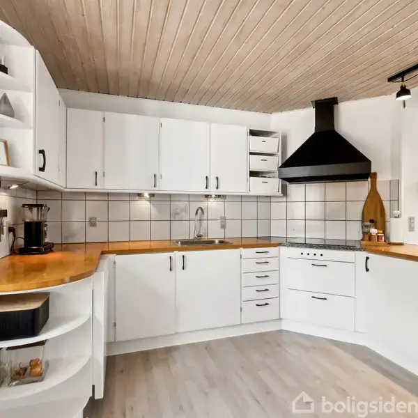 Et moderne køkken med hvide skabe og træbordplader. En kaffemaskine og dekorationer står på bordet. Rummet har gråt flisegulv og et træloft med indbygget belysning.