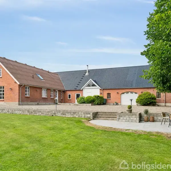 En stor rødstensbygning med skråt tag står i en velplejet have. Foran bygningen er en terrasse med havemøbler. I baggrunden ses en blå himmel.