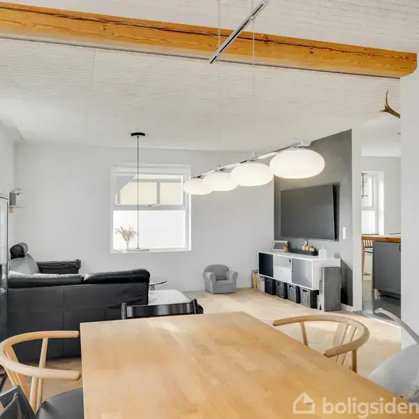 Et moderne, lyst spisestueområde med træbord og stole. Baggrunden har en sofagruppe, en glasskab med porcelæn, og en vægmonteret tv i et hjørne fyldt med naturligt lys.