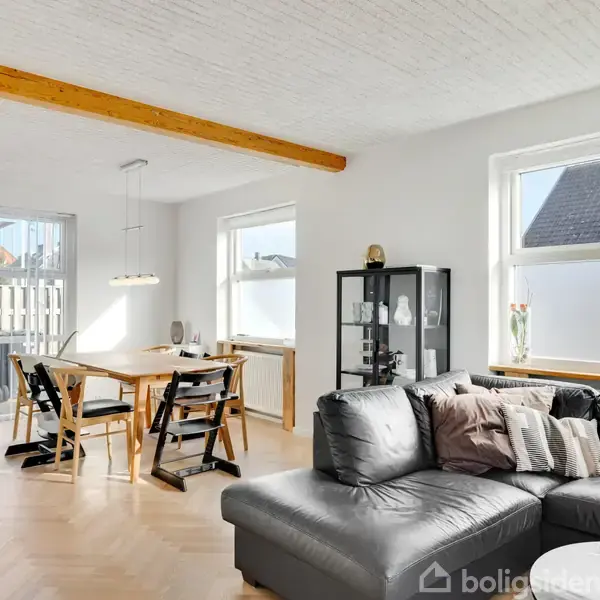 Sofa i stue; står foran vinduer med lysindfald; nær spisebord med stole, glasvitrine, trægulv og hvidmalet loft med en synlig træbjælke.