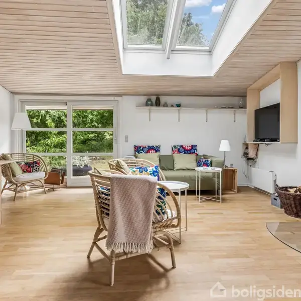 Et lyst stueområde med en brændeovn, komfortable stole og sofa. Store ovenlysvinduer giver naturligt lys. Stuen har træloft og gulv med udsigt over en grøn have gennem glasdøre.