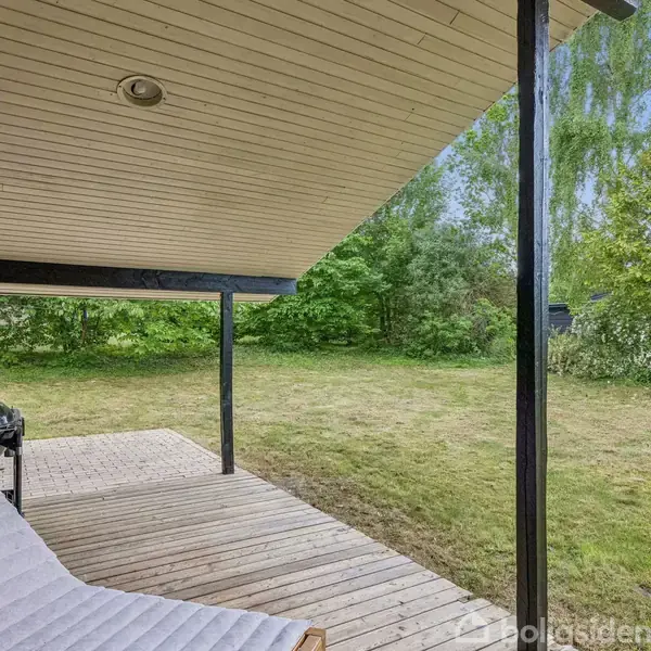 En afslappet solstol står på en træterrasse under et halvtag. Ved siden af er en grill. Terrassen grænser op til en græsplæne med frodige, grønne buske i baggrunden.