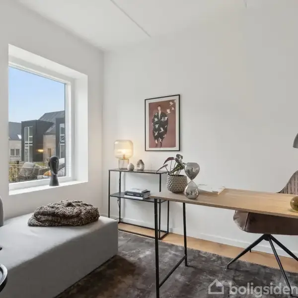 Et lyst kontor med skrivebord og stol, beliggende ved et stort vindue. Bordet har lamper og dekorationer. Væggene prydes med kunst, og rummet indeholder en grå sofa.