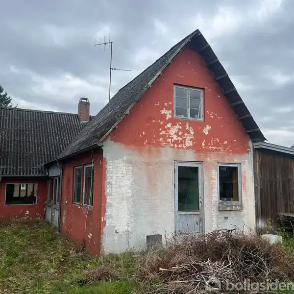 Et gammelt hus med rødmalet, afskallet facade og skiffertag står i en overgroet have. En antenne rager op, og en trædør er placeret centralt.