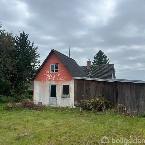 Et ældre hus med rød gavl står stille i en have, omkranset af træer og buske under en overskyet himmel.