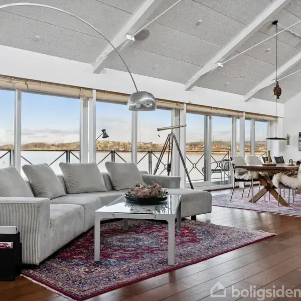 Grå sofa står på et rødt tæppe i en stue med store vinduer, der giver udsigt til et naturskønt område. Et spisebord er til højre, dekoreret med fåreskind.